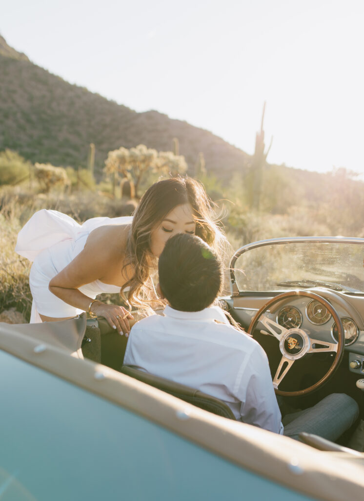 J and Alice's Blue Classic Car Arizona Engagement Session. J sitting inside car A kissing him standing outside of driver's door from behind J