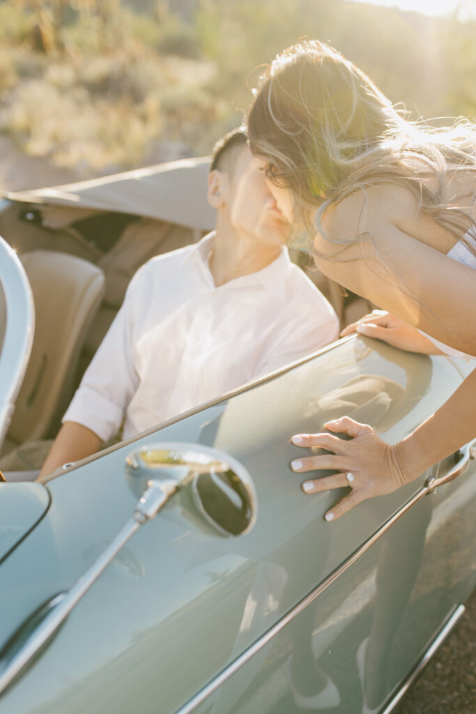 J and Alice's Blue Classic Car Arizona Engagement Session. J sitting inside car A kissing him standing outside of driver's door from behind Alice
