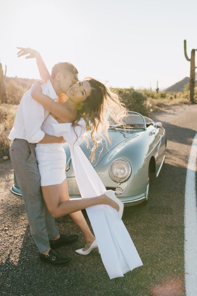 J and Alice's Blue Classic Car Arizona sunrise Engagement Session. J & A standing in front of the blue classic car sharing a passionate embrace and laughter