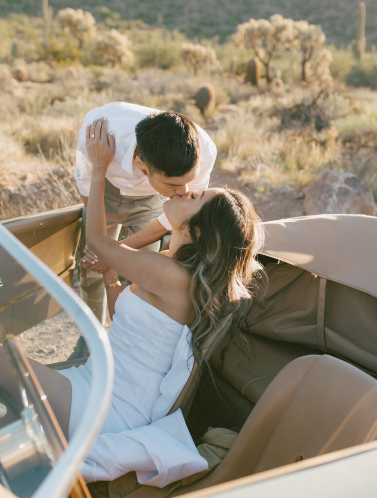 J and Alice's Blue Classic Car Arizona Engagement Session. A sitting inside car J kissing her standing outside of driver's door from behind A