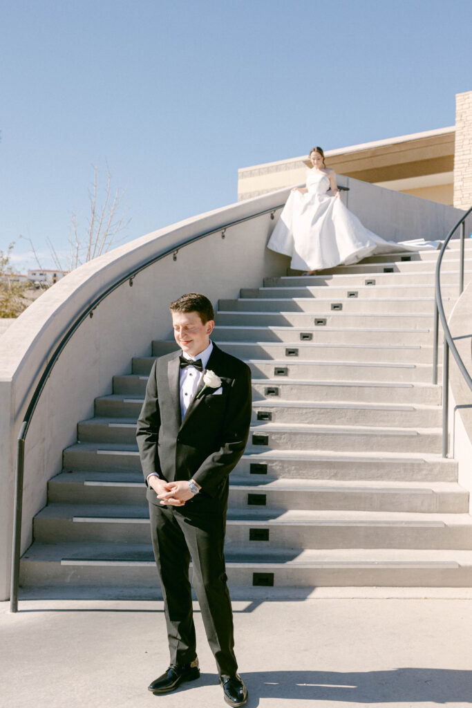 A groom in a sharp black tuxedo standing at the base of a grand staircase, with the bride gracefully posing at the top under a clear blue sky.