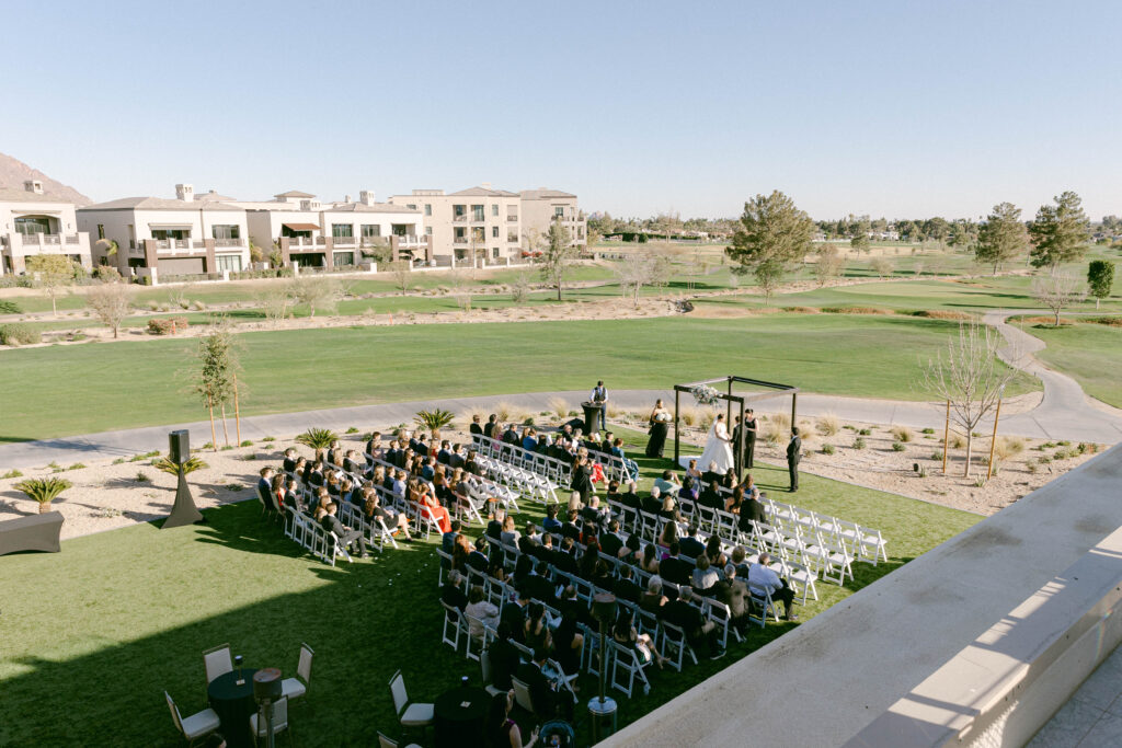 Outdoor wedding ceremony at the Arizona Biltmore Golf Club with a scenic golf course and mountain views in the background. Guests are seated on a manicured lawn as the couple exchanges vows under a modern black ceremony arch.