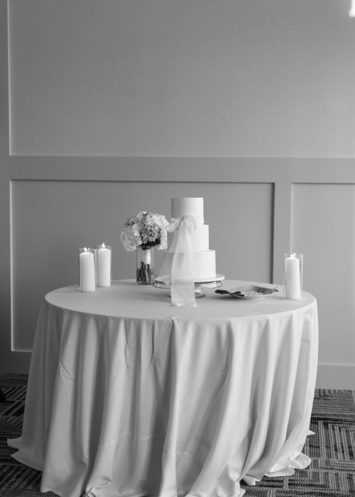 A three-tiered white wedding cake adorned with a delicate tied bow, elegantly displayed on a table in the Arizona Biltmore Golf Club ballroom.