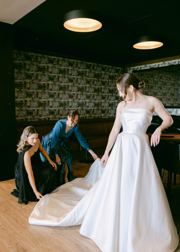 A bride in a sleek, strapless wedding gown having the final touches done to her dress, with bridesmaids assisting in an elegant indoor setting.