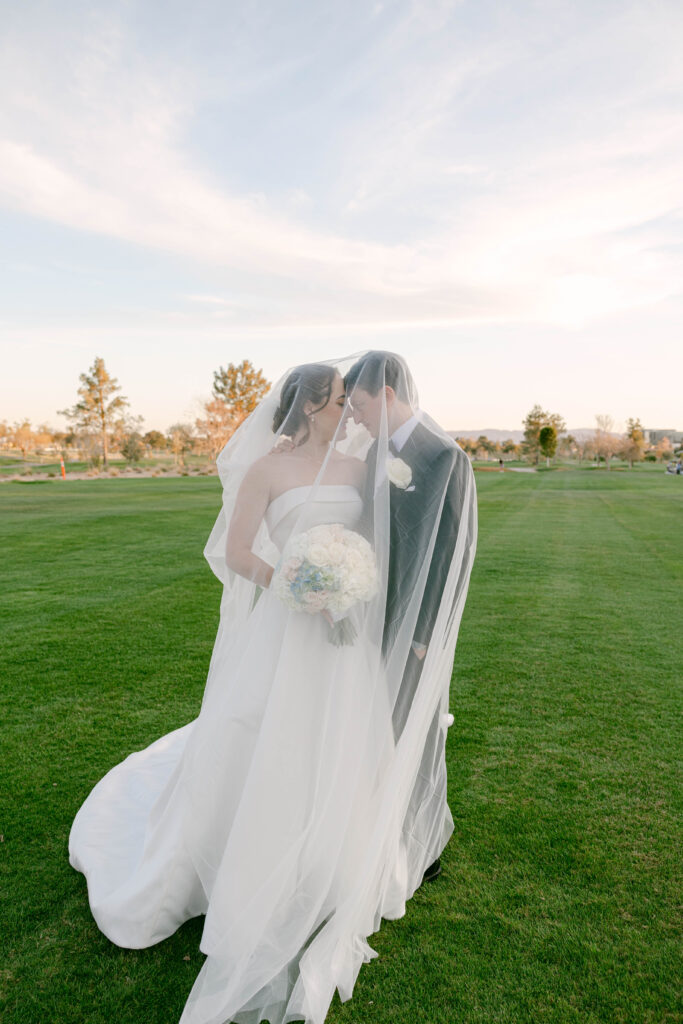 Bride in a flowing white gown with a long veil, walking on the green lawn of the Arizona Biltmore Golf Club at sunset.