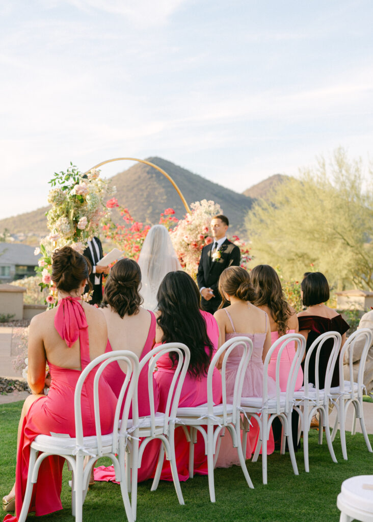 Bridesmaids in mix-and-match pink dresses seated during the wedding ceremony, watching the bride and groom exchange vows with joy and admiration.